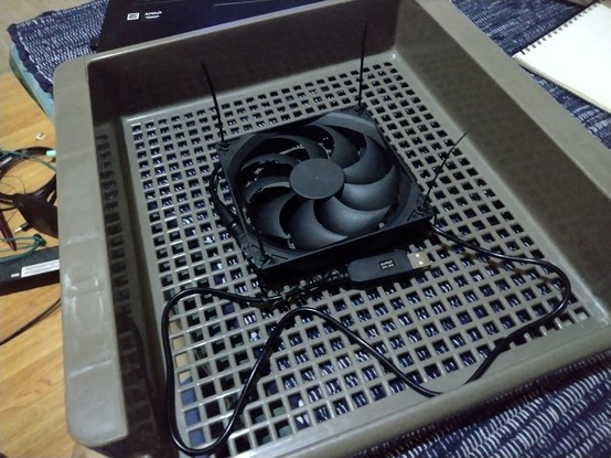 Underside of a DIY laptop cooling pad. The base is a part of a plastic shelf. The computer fan is fixed at the middlw of the base with zipties on each corner. Connected to the fan is a USB cable that steps up 5v input into 9v output.