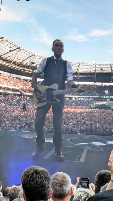 Bruce Springsteen live auf der Bühne. aufgenommen vom großen Bildschirm. Bruce im Vordergrund und das Stadion voller Besucher im Hintergrund.