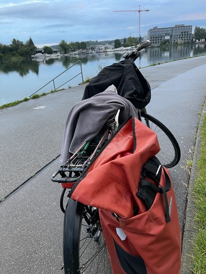 Ein Fahrrad mit roter Satteltasche. Es steht an einem Fuß- und Fahrradweg auf dessen linker Seite der Seerhein ist und auf dessen rechter Seite sich eine Wiese befindet. Auf dem Sattel liegt ein Handtuch, auf dem Lenker eine Regenjacke 