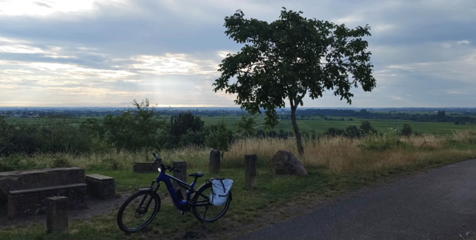 Auf einer Anhöhe über der weiten Rheinebene steht eine Sitzgruppe aus Sandstein an einem Feldweg. Ein Nussbaum wächst daneben. Mein Fahrrad steht davor. Unten in der Ebene sieht man endlose Weinpflanzungen. Der Himmel ist größtenteils bewölkt.