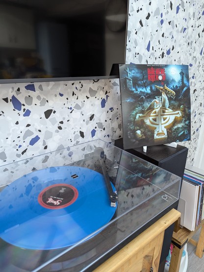 Blue vinyl of Rite Here Rite Now playing on a turntable with the album cover standing upright next to it.