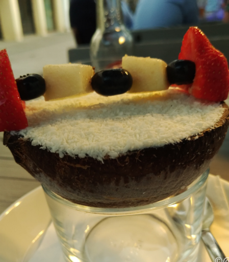 Auf einem Glas sitzt eine halbe Kokosnussschale, gefüllt mit Kokoseis, oben drauf Kokosraspel und ein Obst-Spieß mit 2 Erdbeerhälften, 3 Blaubeeren und 2 Melonenstückchen.