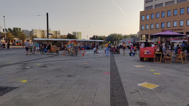 Ein Überblick über die Westseite des Alice Salmon Platzes in Hellersdorf in Richtung der U-Bahn. Es sind einige Stände zu sehen, ein bunter Regenbogen-Fahrradanhänger, ein 