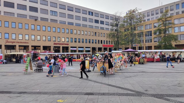 Eine weitere Totalaufnahme des westlichen Alice Salmon Platzes, diesmal in Richtung des Rathauses. Wieder zu sehen die zahlreichen Stände, die Lastenräder und Anbringungen auf dem Boden. Im Hintergrund sieht man die Geschäfte wie Bäckerei Junge, Woolworth, etc.