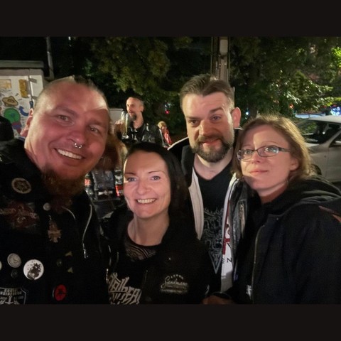 Selfie von 4 Personen, die in der Konzertpause vor der Halle stehen, um frische Luft zu schnappen. Im Hintergrund erkennt man verschwommen weitere Konzertbesucher, parkierende PKW und Bäume. 