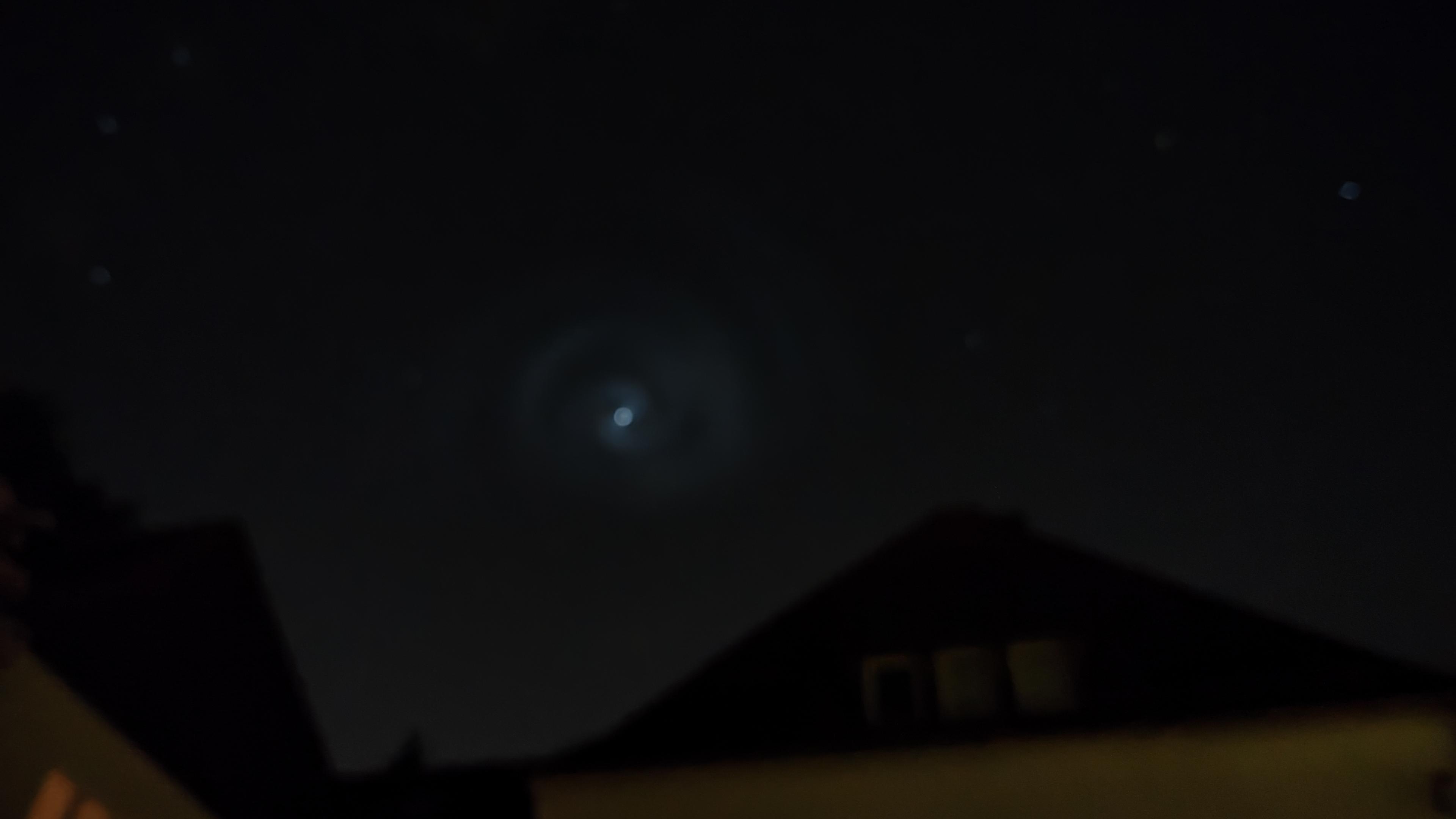 Der Nachthimmel über einen Hausdach. Ein Stern oder etwas in der Größe eines Sterns ist umgeben von einer ganz blassen, aber deutlichen Lichtspirale. Kein gewöhnlicher Anblick. 