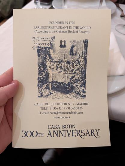 The image shows a card held by a hand, featuring information about Casa Botín, the earliest restaurant in the world according to the Guinness Book of Records, founded in 1725. The card is cream-colored with black text and illustrations. At the top, it states "FOUNDED IN 1725 EARLIEST RESTAURANT IN THE WORLD (According to the Guinness Book of Records)." Below this, there is an illustration of a dining scene with people seated around a table, and a server in an apron. The address "CALLE DE CUCHIL…