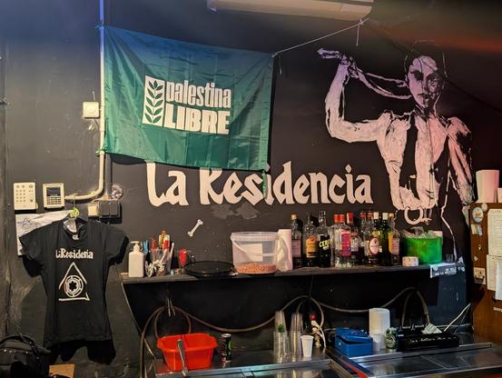 The image depicts an interior space, likely a bar or a small venue, with a dark wall as the backdrop. On the left side, a green flag with the text "palestina LIBRE" is prominently displayed. Below the flag, a black T-shirt with the text "La Residencia" hangs on the wall. The central part of the wall features a large white text "La Residencia" and a mural of a person with a hand behind their head, wearing a suit and tie. The right side of the wall has a shelf with various bottles of alcohol, inc…