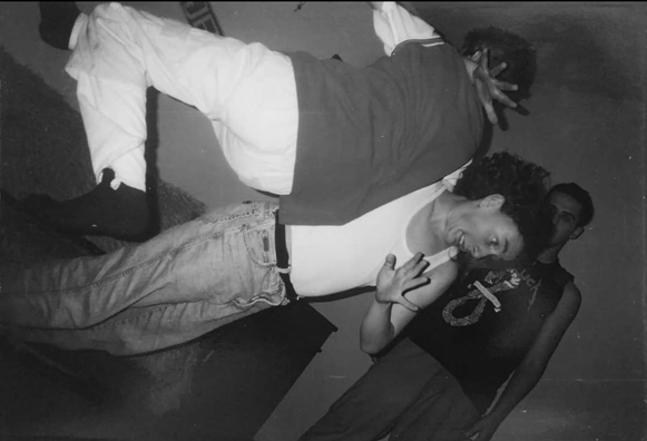 The image is a black and white photograph featuring three individuals in a dynamic pose. The central figure is leaning back with their head tilted to the side, wearing a white sleeveless shirt and light blue jeans. They are making a hand gesture near their face. The person behind them is wearing a dark shirt with a graphic design and dark pants, with their hand raised. The third person is partially visible, wearing a dark shirt and pants, standing behind the central figure. The background is pl…