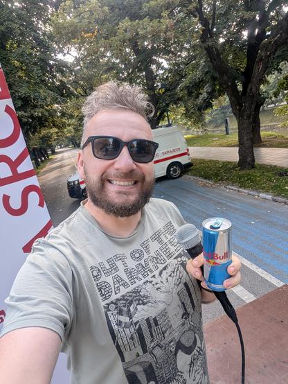 The image shows a man taking a selfie on a sidewalk. He is wearing a light gray t-shirt with a graphic design that includes the text "OUT Of THE DARKNESS" and an illustration of a cityscape. The man is holding a Red Bull can in his right hand and a microphone in his left hand, with a cable attached to the microphone. He is wearing black sunglasses and has a beard. In the background, there is a white ambulance van with red text that reads "SARAJEVO," parked on the side of a road with a blue bike…