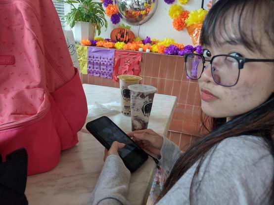 My GF holding her phone, looking at the camera from her side. On the table are two bubble milk tea. In the background are Day of the Dead decorations