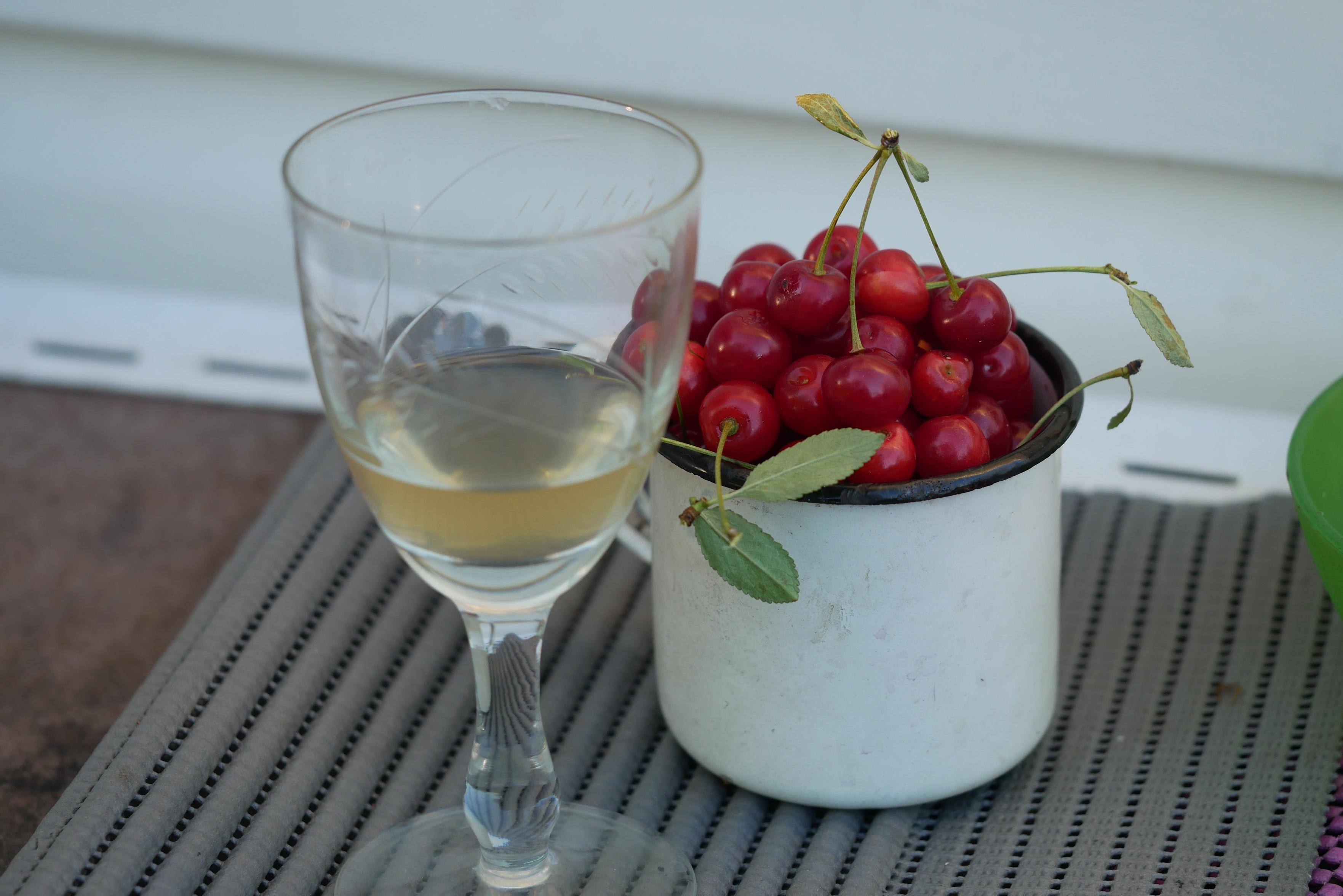 On a table near the wall: an old cracked glass glass with a yellow liquid (mead - honey drink), next to a white enameled mug filled with cherries.