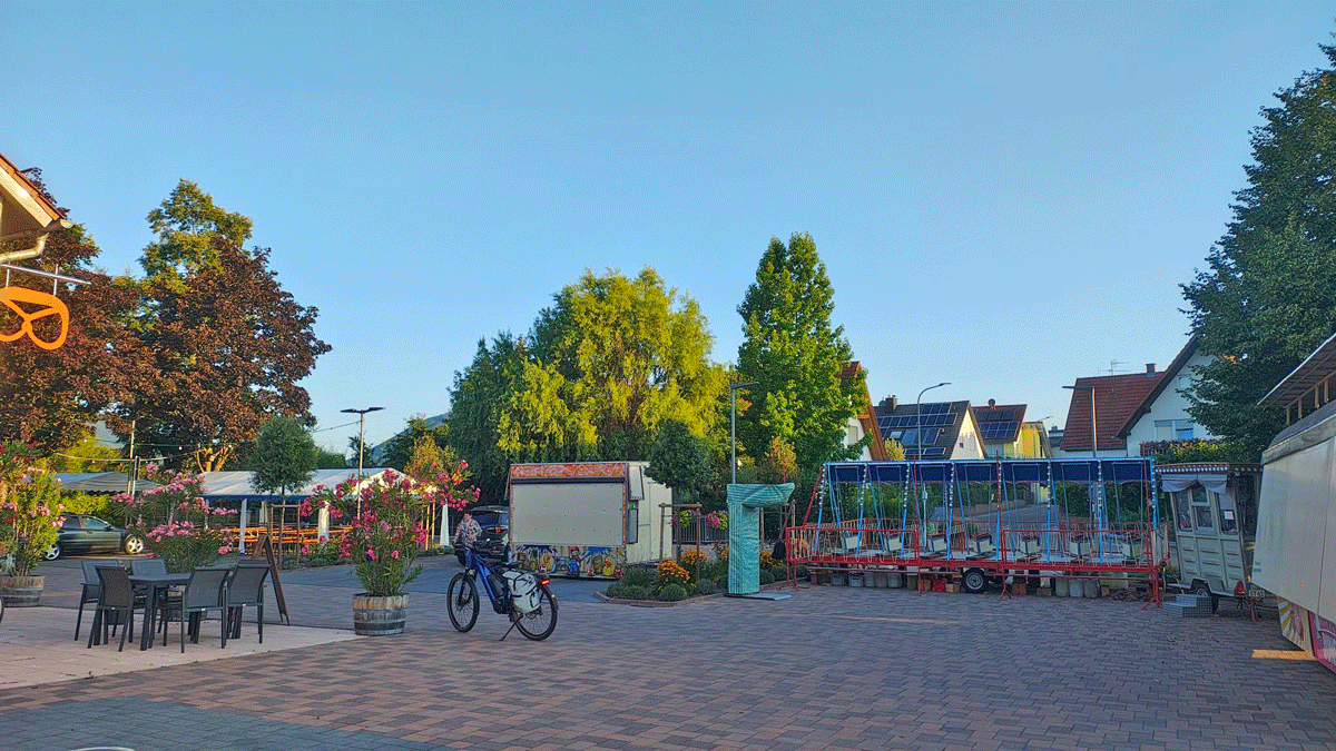 Ein Dorfplatz am Morgen. Links ein Gebäude mit an der Fassade aufgehängter Brezel. Hier kaufe ich manchmal meine Brötchen morgens auf dem Weg zur Arbeit. Daneben stehen ein Weinzelt, so eine Box-Gerätschaft für Jahrmärkte und ein Süßigkeitenstand, alle verschlossen. Dazwischen ist eine alte Schiffschaukel aufgebaut, mit sieben kleinen Schaukeln in Bootsform, die man mit eigener Muskelkraft antreiben muss.
