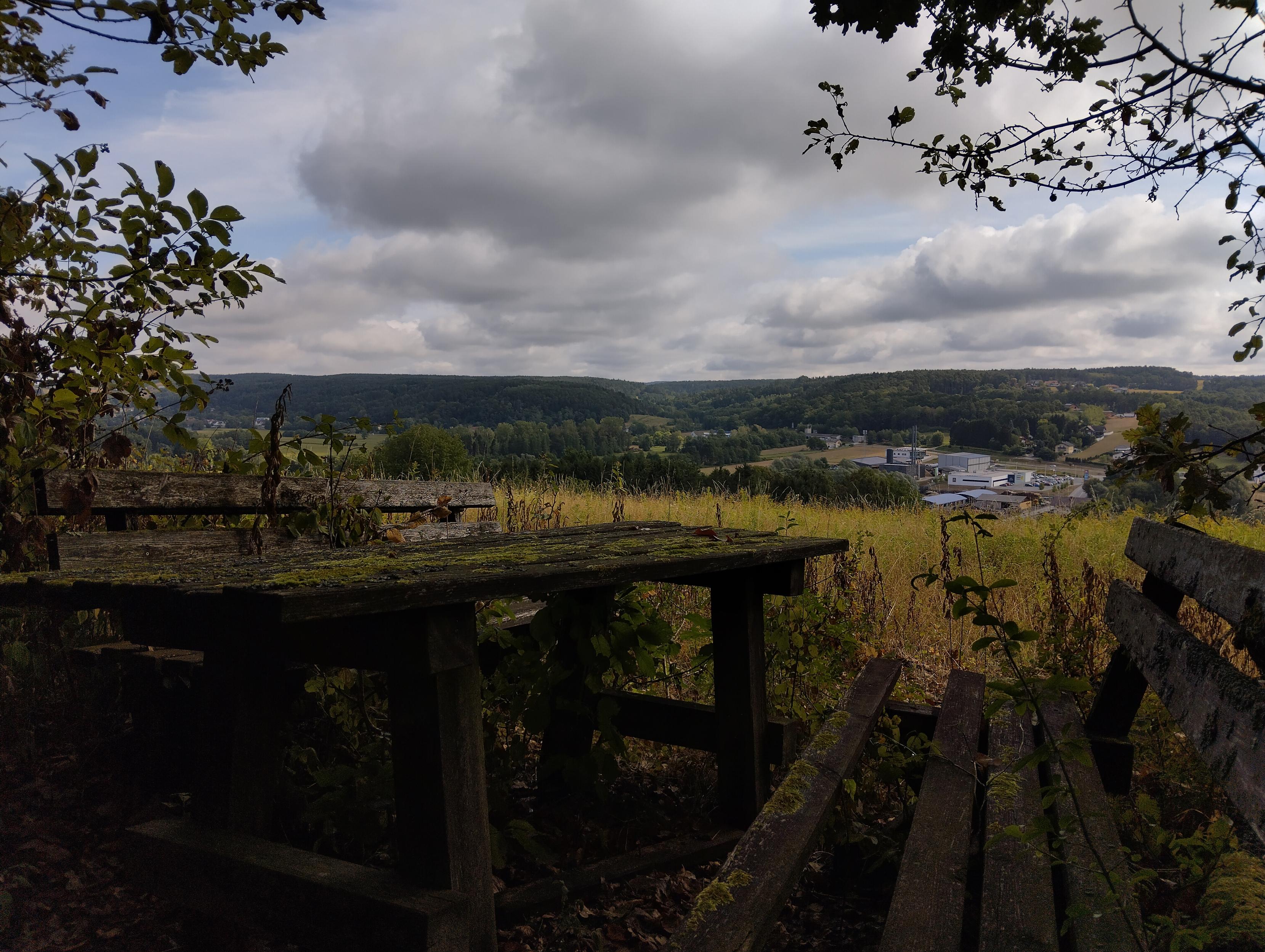 photo depicts two wooden benches and a wooden table between them, the wood itself is old, rotten and covered moss, planks are missing here and there, plants grow between and through the planks, smal trees shade the spot from the sides, behind the rest spot a gentle slope with soft green vegetation gives way to a valley with a village and dark green forests on the hills beyond 
the sky is covered in grey-white clouds and occasional blue sky